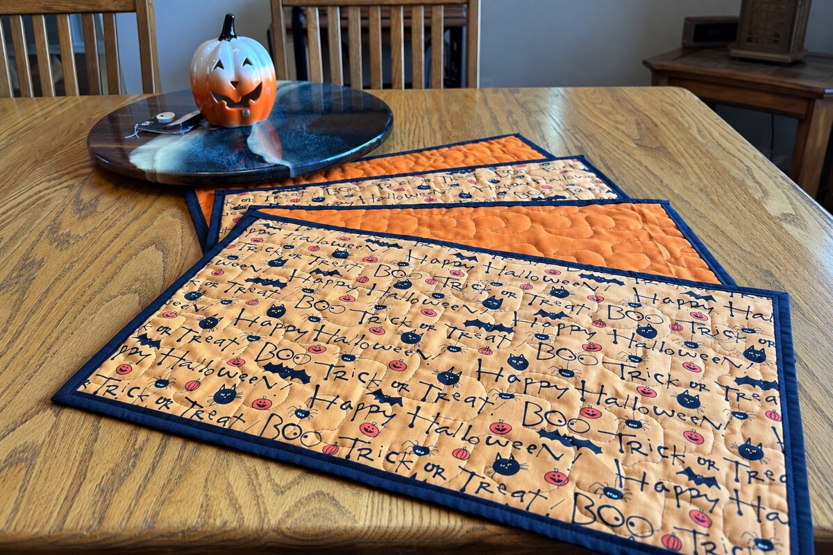 Four finished longarm-quilted Halloween placemats with black binding displayed beside a ceramic pumpkin